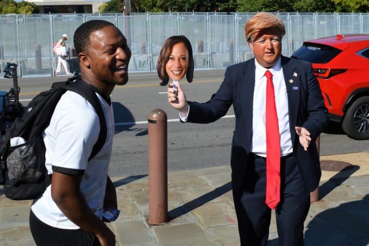 An imitator dressed as Donald Trump waves a picture of Kamala Harris on a stick, while a man standing closely to the imitator laughs.