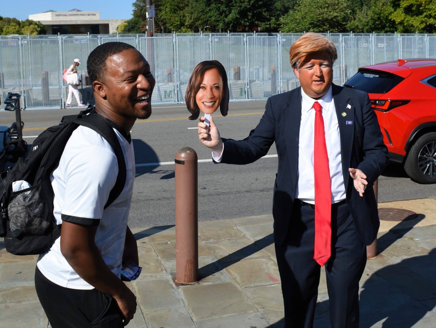 An imitator dressed as Donald Trump waves a picture of Kamala Harris on a stick, while a man standing closely to the imitator laughs.