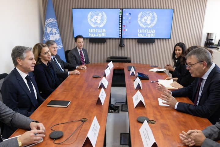 A conference table around on either side of which sit many diplomats and aid workers; chiefly U.S. Secretary of State Anthony Blinken on one side and UNRWA Commissioner-General Philippe Lazzarini on the other, who is speaking. Behind them are two screens displaying the UNRWA logo.