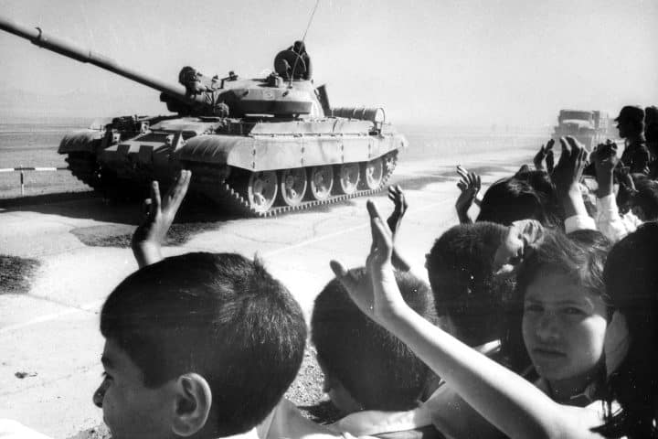 A tank going down a road with children waving after it.