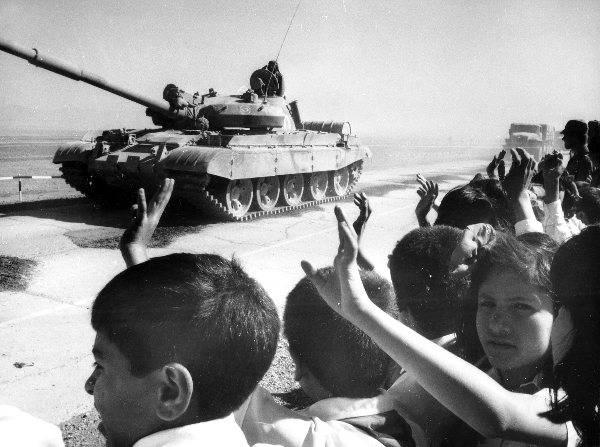 A tank going down a road with children waving after it.