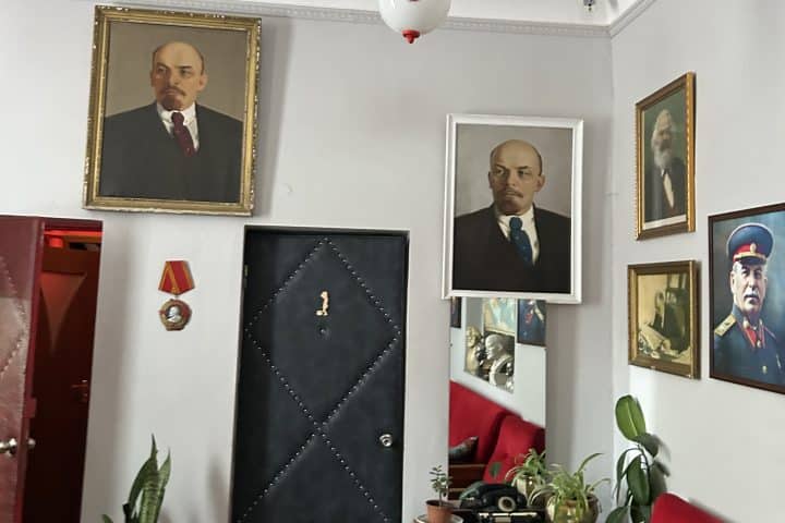 A room which has several paintings of communist figures, prominently Lenin but also Stalin and Marx. Vases with plants are scattered around.