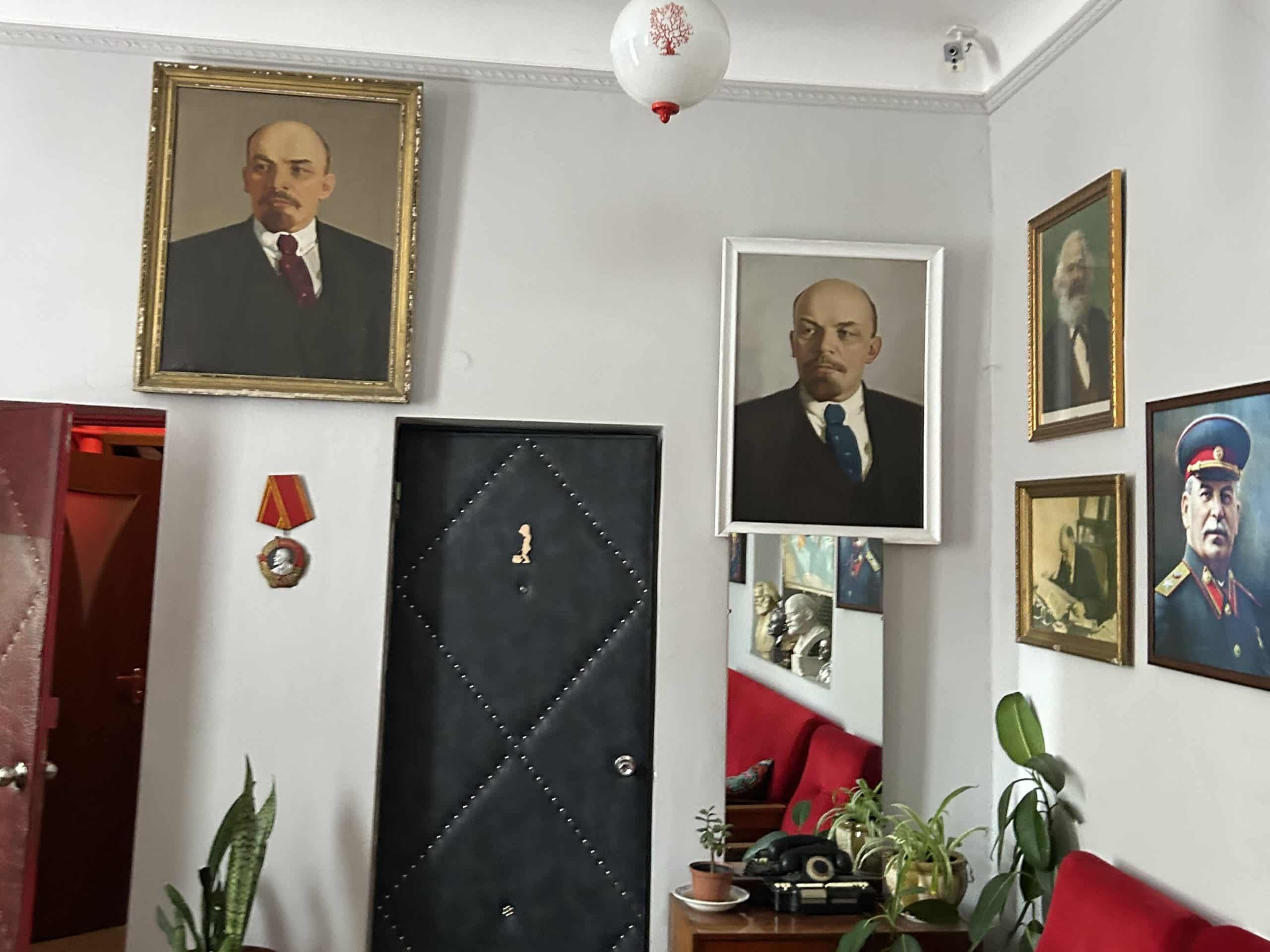 A room which has several paintings of communist figures, prominently Lenin but also Stalin and Marx. Vases with plants are scattered around.