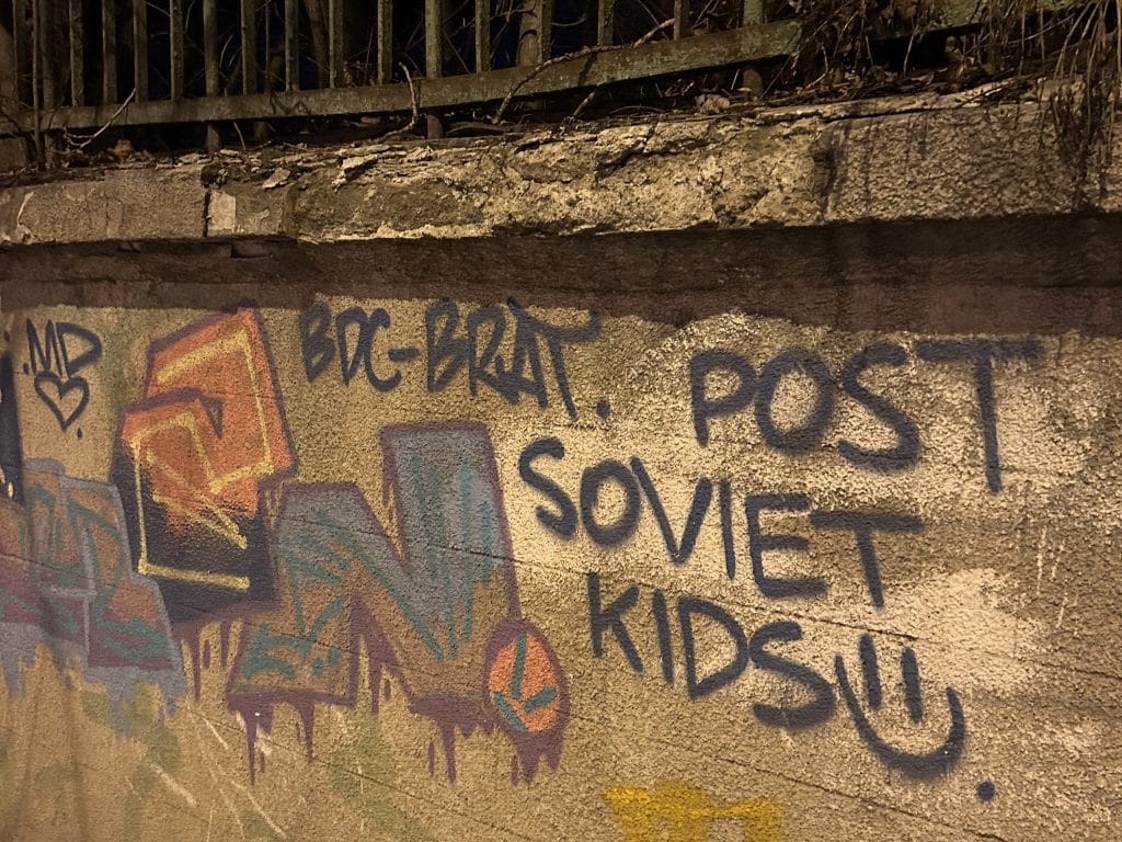 A concrete barrier with graffiti on it, reading: "POST SOVIET KIDS", followed by a smiley.