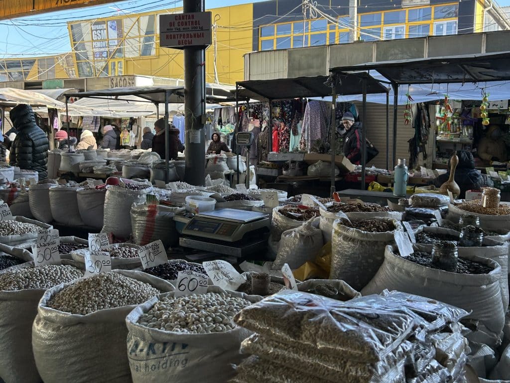 Bags of seeds, vegetables and other foodstuffs line an outdoor market. People walk in the background.