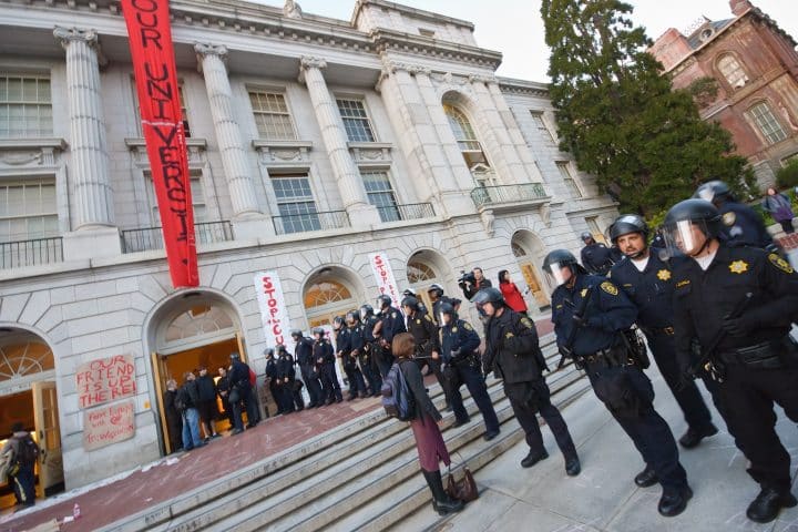 A university building, with police officers in riot gear standing in a line beside it.